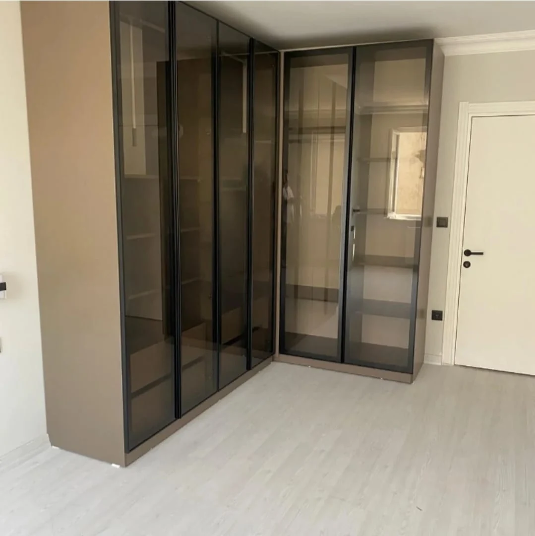 Cam kapaklı köşe giyinme alanı. Köşeyi çevreleyen koyu çerçeveli cam kapakların ardında raf ve askı bölmeleri görülüyor.